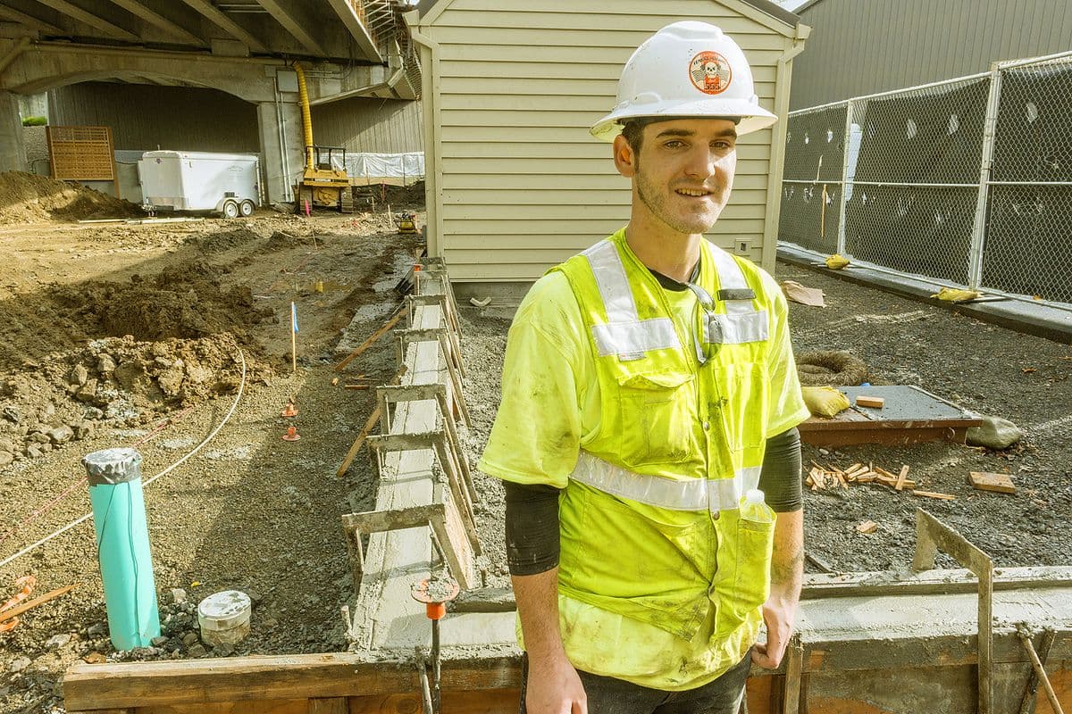 A cement mason apprentice in a hard hat and high-visibility shirt stands beside fresh concrete forms on an Oregon job site.