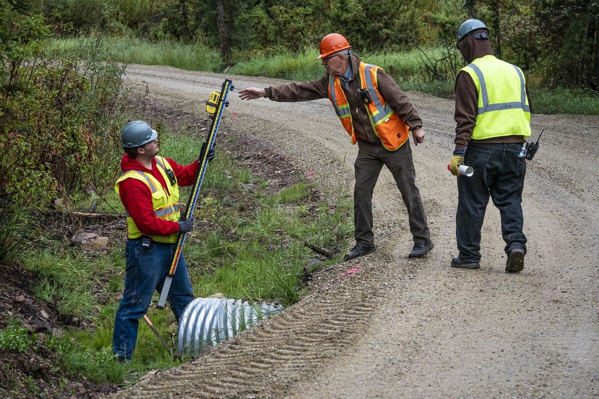 A trainee holds a measuring rod next to a culvert while an instructor points down a gravel road and another trainee watches.
