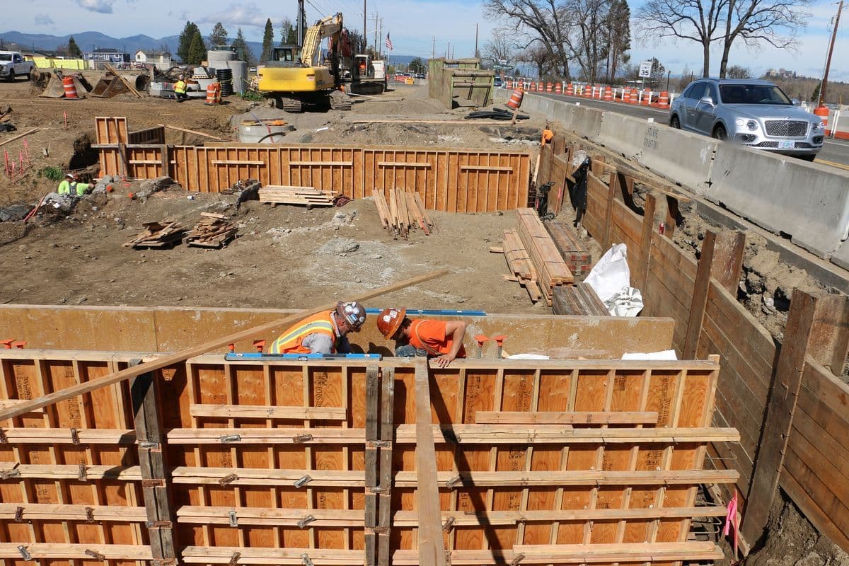 Two workers in hard hats check wooden concrete forms for new bridge supports beside a highway in southern Oregon.