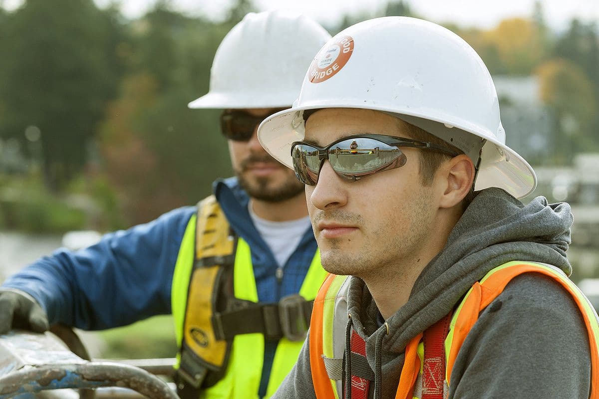 An electrician apprentice in a hard hat and safety glasses on a bridge project in Oregon, with a coworker behind him.